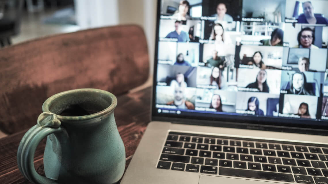 Foto eines Laptops auf dem eine Videokonferenz zu sehen ist. Daneben steht eine Tasse Kaffee