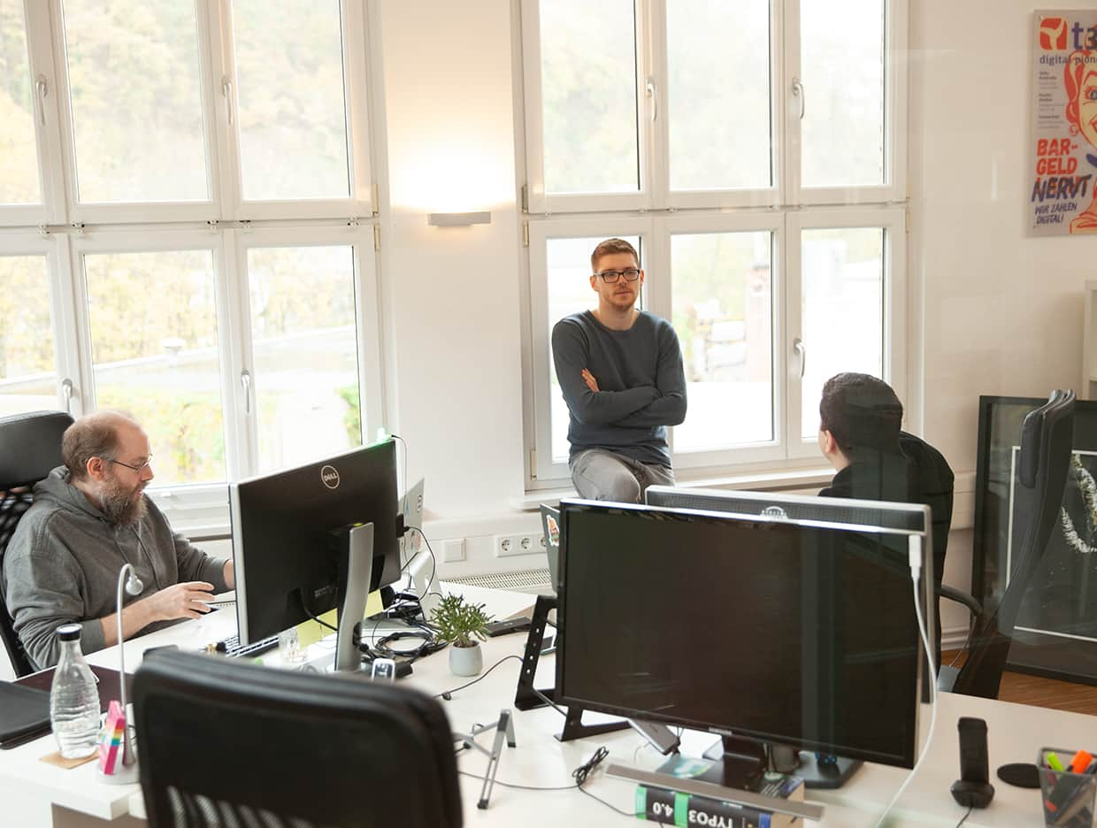 Foto einer Büroecke mit 3 Arbeitsplätzen. Daran sitzen Daniel Phillips und Daniel Schultheis und besprechen etwas mit Eric Groß welcher am Fenster steht